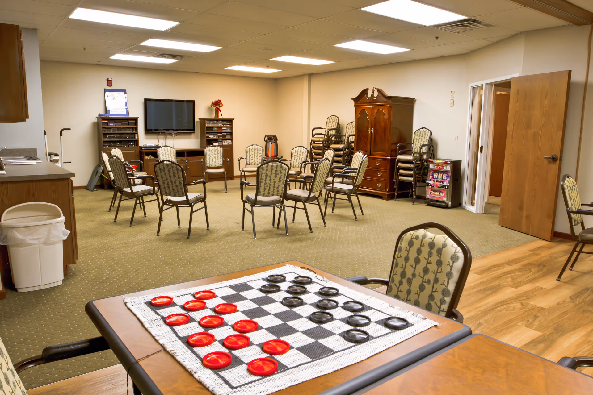 A spacious activity room in a senior living facility with a checkers game set up on a table in the foreground. The room has multiple chairs arranged in a circle, a television mounted on the wall, wooden cabinets, and a carpeted floor. There is a wooden armoire and a slot machine in the corner, with an open door leading to another room.