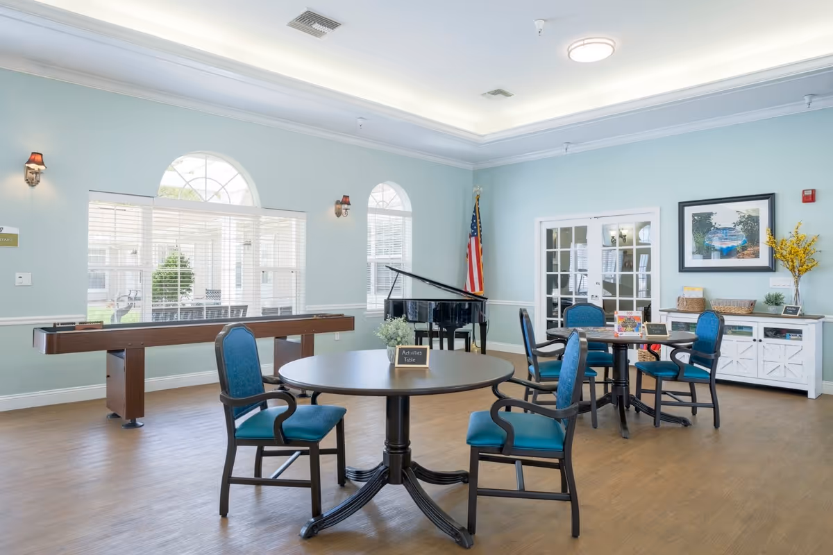 A bright and spacious activity room with light blue walls and wood flooring. The room features round tables with blue cushioned chairs, a shuffleboard table, a black grand piano, and an American flag in the corner. Large windows with white blinds allow natural light to fill the space. A white sideboard with decorative items and a framed picture hangs on the wall.