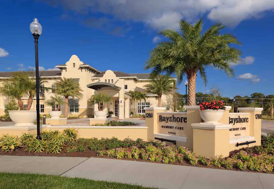 Front exterior of the Bayshore Memory Care building showing the entrance, signage, palm trees, and landscaped grounds.