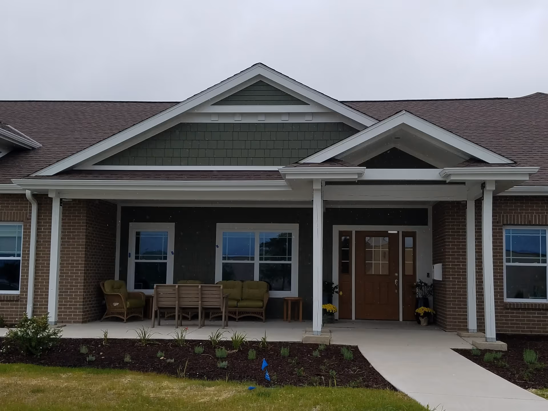 Front exterior view of a single-story building with a covered porch area featuring outdoor seating including chairs and a small table. The building has a brown shingled roof, green siding with white trim, and brick walls. There are windows on either side of a wooden front door with glass panels. The walkway leads up to the entrance, and there are small plants and mulch beds in front of the porch.
