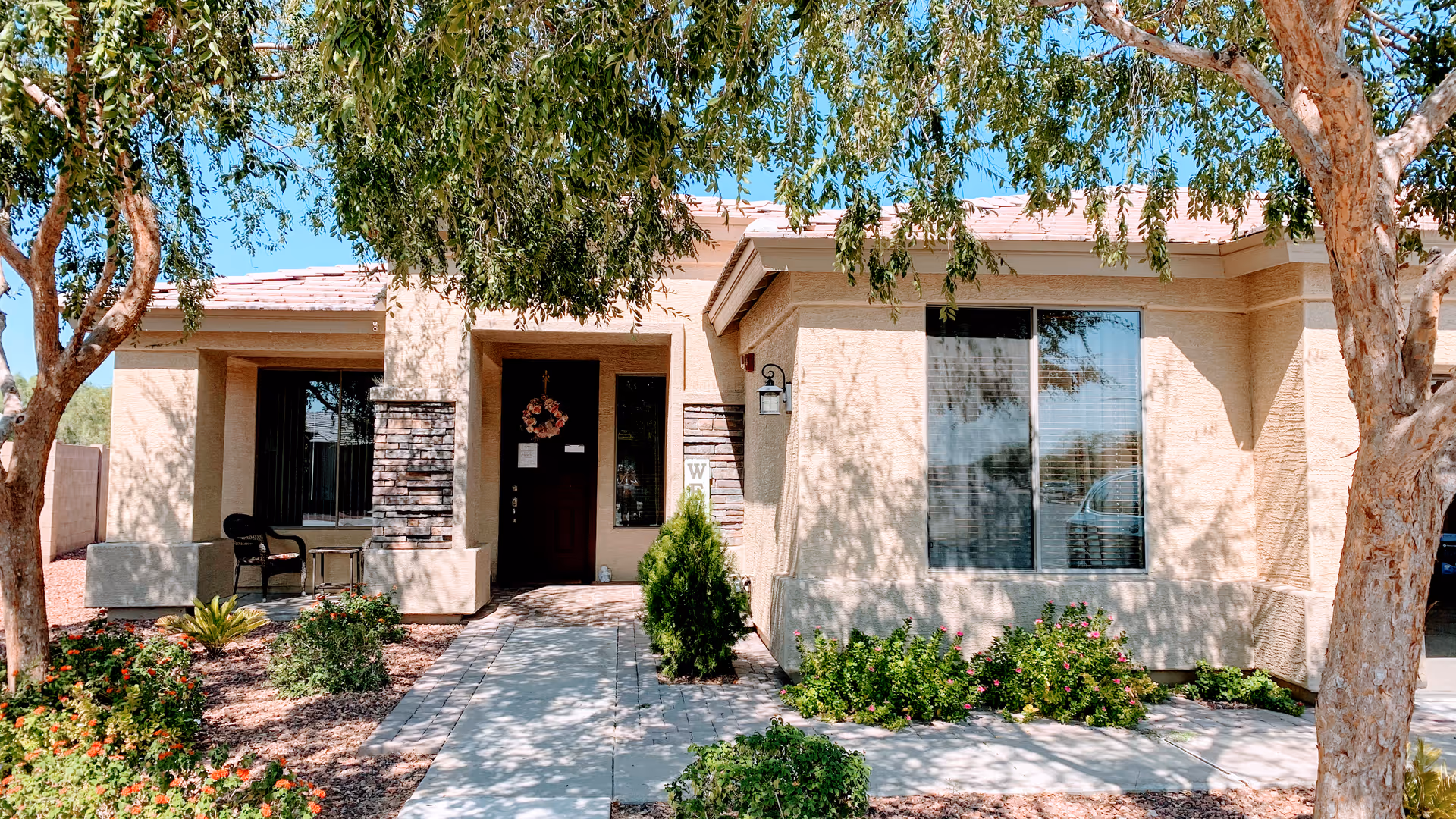 Front exterior view of a single-story residential building with a covered entrance, a pathway leading to the front door, and landscaping including trees and bushes under a clear blue sky.