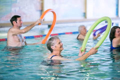 A group of elderly people participating in a water exercise class in an indoor swimming pool, each holding colorful pool noodles and smiling.