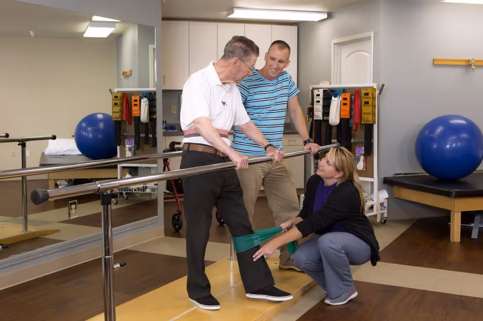 An elderly man is performing a physical therapy exercise using parallel bars while a female therapist kneels and assists by holding a resistance band around his legs. A male therapist stands nearby, observing and supporting the session in a rehabilitation room equipped with exercise balls and therapy equipment.
