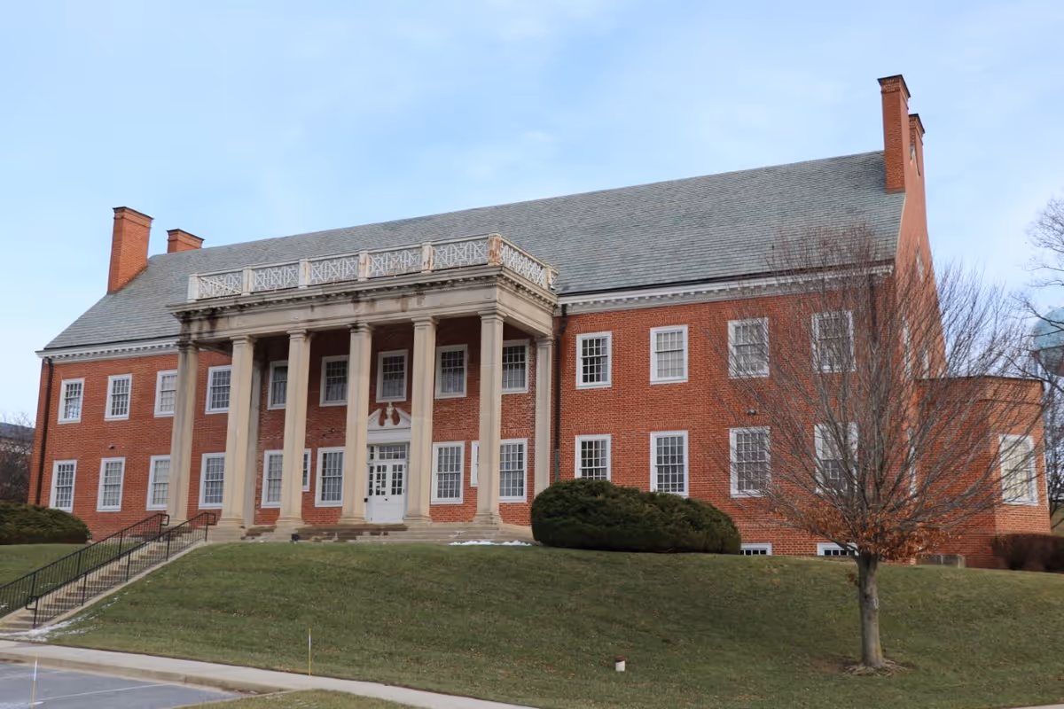 A large, historic red brick building with white-framed windows and tall columns at the entrance. The building has a sloped gray roof with chimneys and is surrounded by a grassy lawn with a few bushes and a leafless tree. The sky is clear and blue.