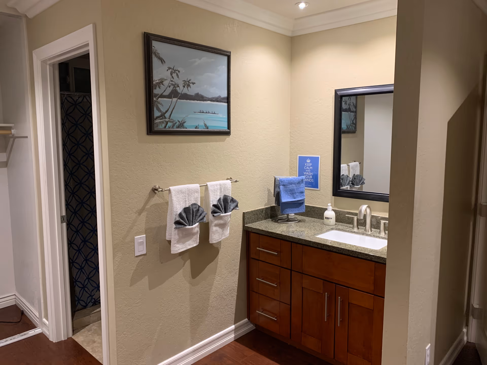 A bathroom vanity area with a granite countertop, a sink, and a mirror above it. Two white towels with decorative gray folded towels are hanging on a towel rack on the wall. A blue towel is hanging on a stand next to the sink. A framed painting of a tropical beach scene is hung on the wall above the towel rack. The bathroom entrance with a shower curtain featuring a blue circular pattern is visible to the left.