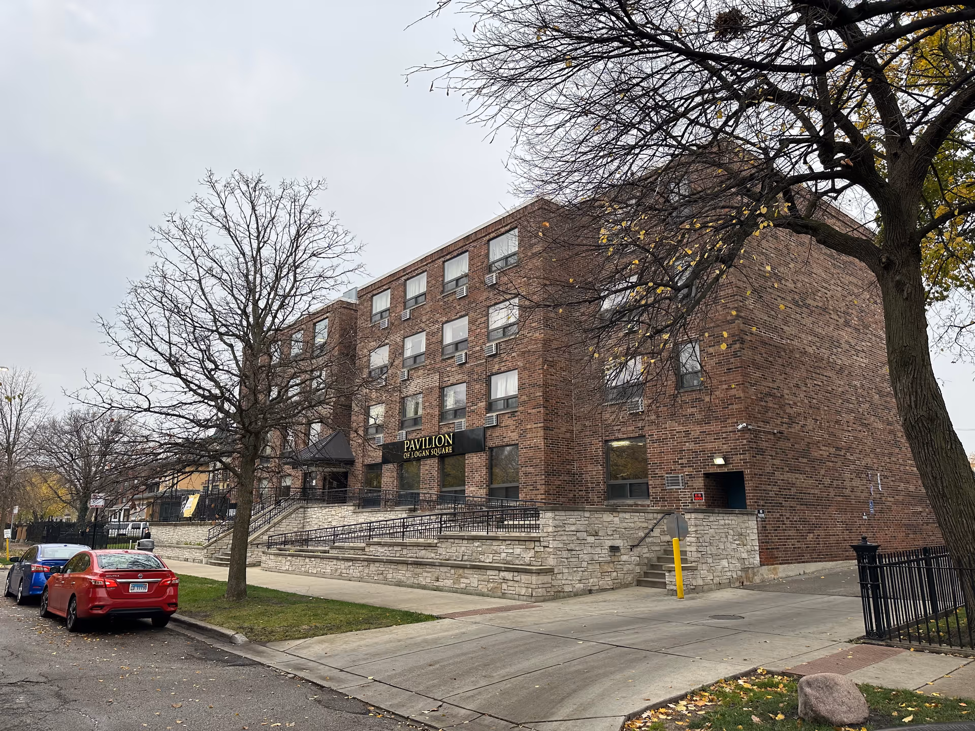 Exterior view of a four-story brick building named Pavilion of Logan Square with a stone base and a ramp leading to the entrance. There are leafless trees and parked cars along the street in front of the building under an overcast sky.