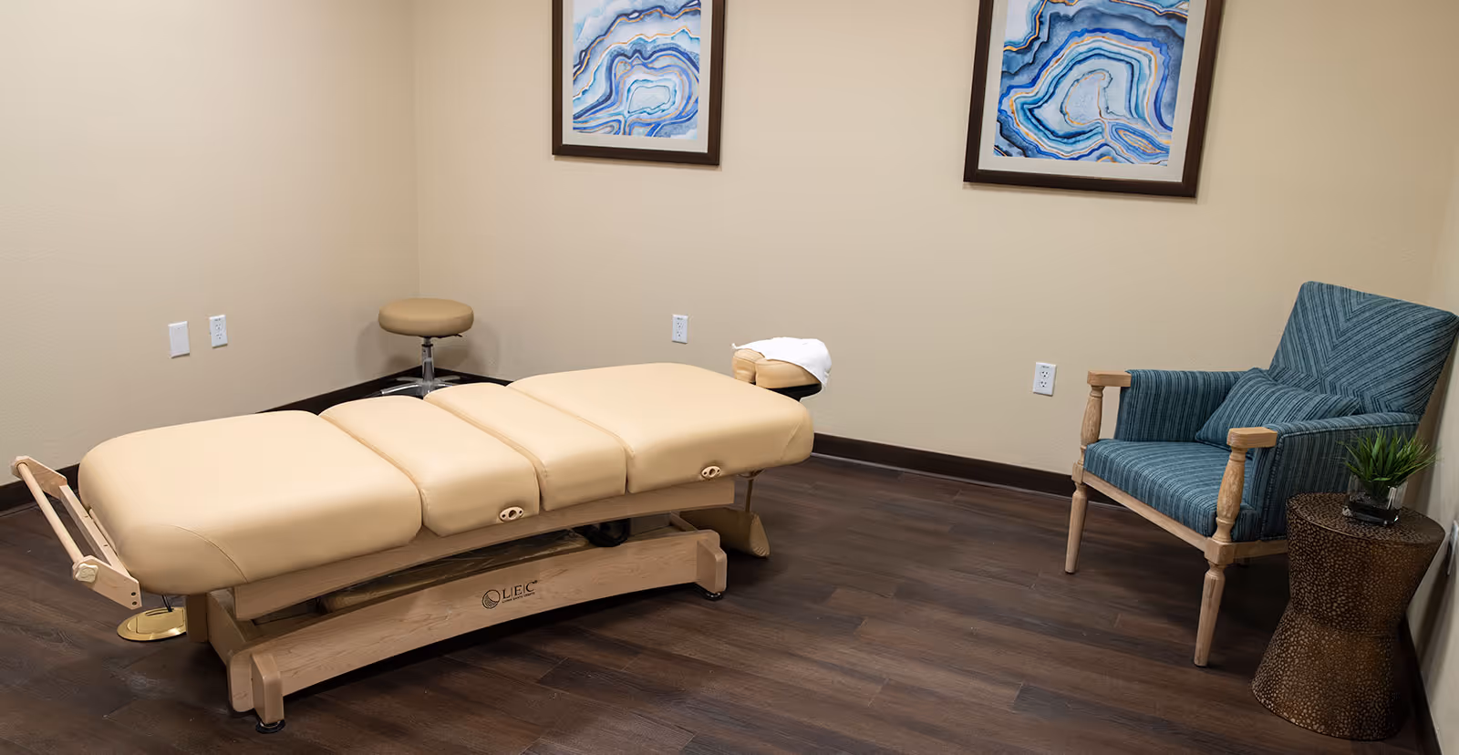 A therapy or massage room with a beige padded treatment table, a small beige stool, a blue upholstered armchair with a matching pillow, a small round side table with a plant, and two abstract blue and white framed artworks on the wall.