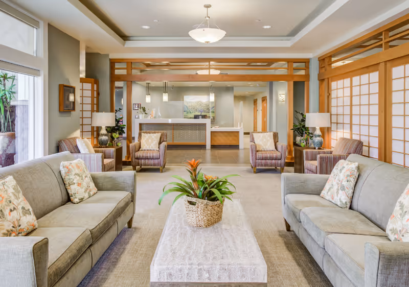 A bright and spacious senior living facility lounge area with two gray sofas facing each other, each adorned with floral patterned pillows. Between the sofas is a rectangular coffee table with a woven basket containing green plants and orange flowers. In the background, there are four armchairs with striped upholstery and a reception desk with a landscape painting on the wall behind it. The room features wooden accents, large windows with white blinds, and soft lighting from ceiling fixtures and table lamps.