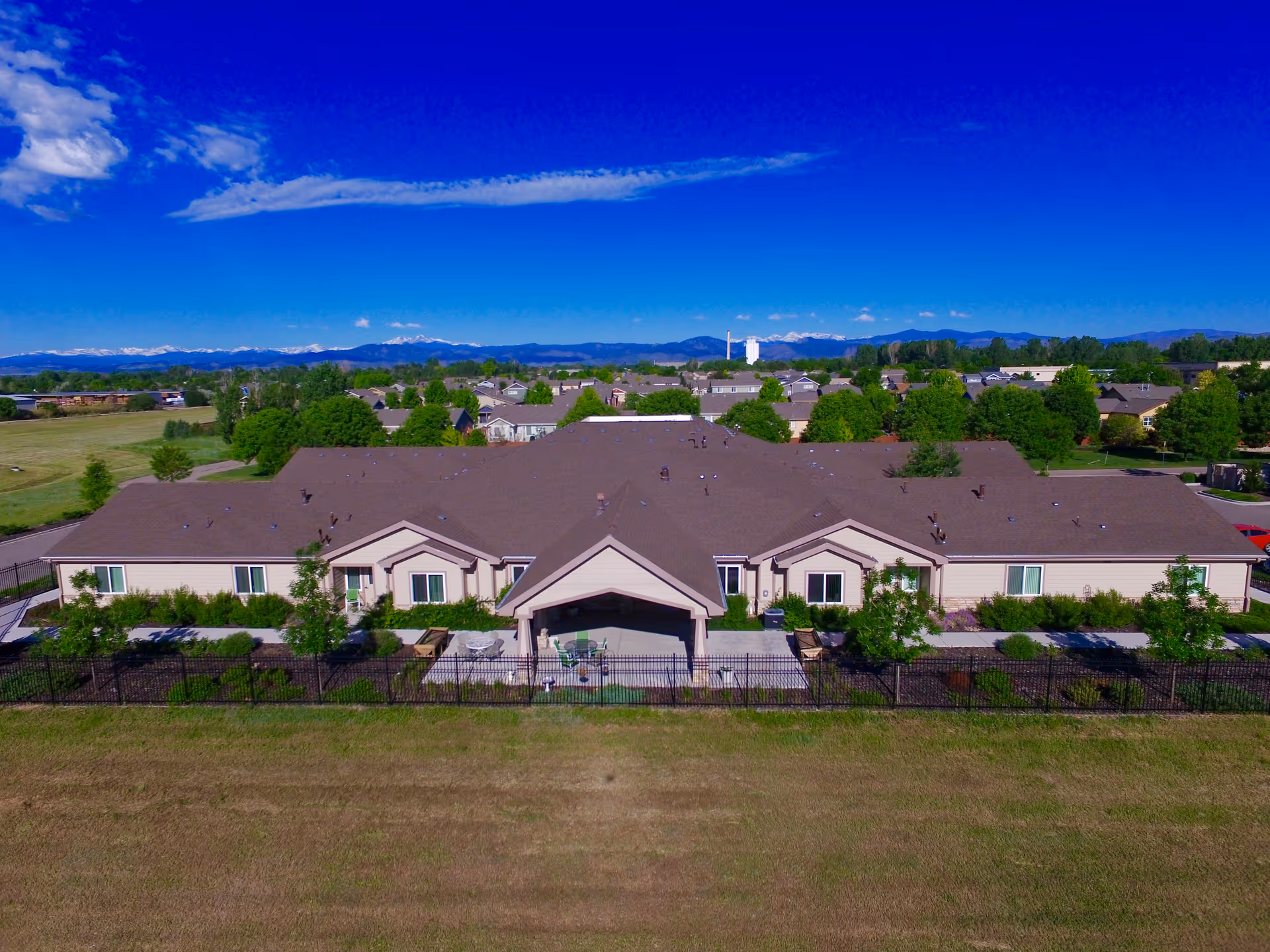 Aerial view of a single-story senior living facility building with a brown roof and beige exterior walls, surrounded by a black fence and greenery. The facility is set against a backdrop of a suburban neighborhood with trees and distant mountains under a clear blue sky.