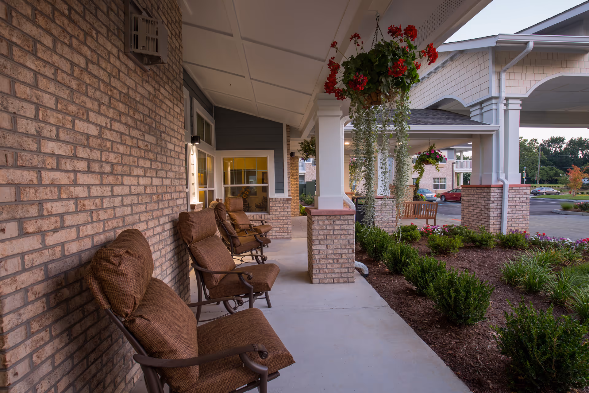Covered outdoor patio area at Willow Creek Gracious Retirement Living with cushioned brown chairs lined up against a brick wall, hanging flower baskets, and landscaped garden beds along the walkway.