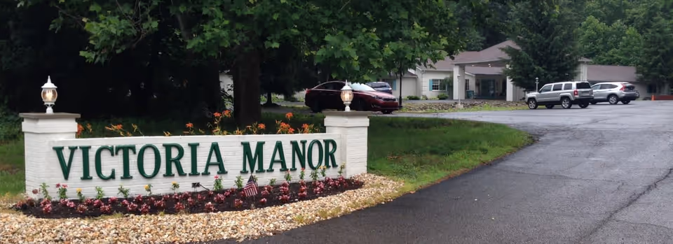 Entrance sign for Victoria Manor with green lettering on a white brick structure, surrounded by flowers and landscaping. In the background, there is a driveway leading to a building with several parked cars and trees around.