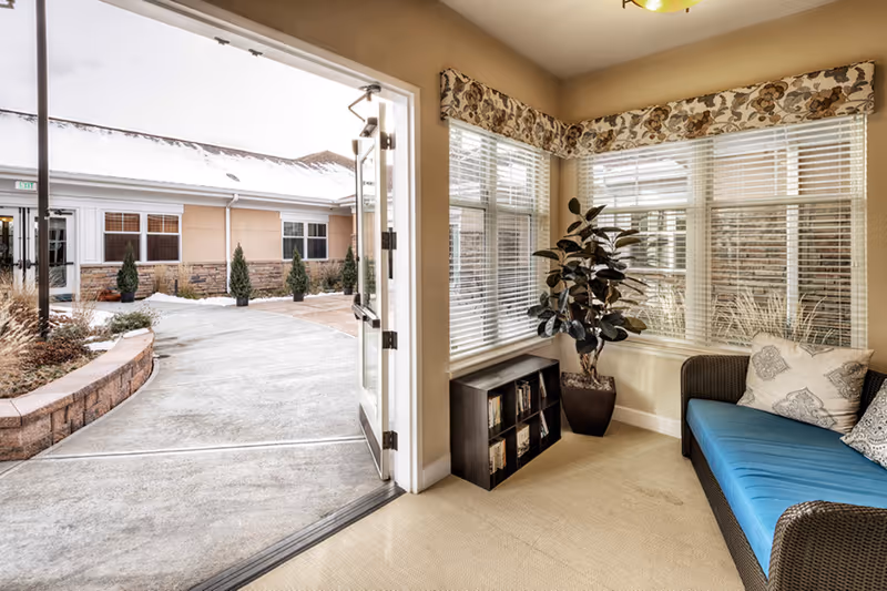 Sunny seating area with a blue couch, potted plant and bookshelf beside an open door leading to a courtyard.