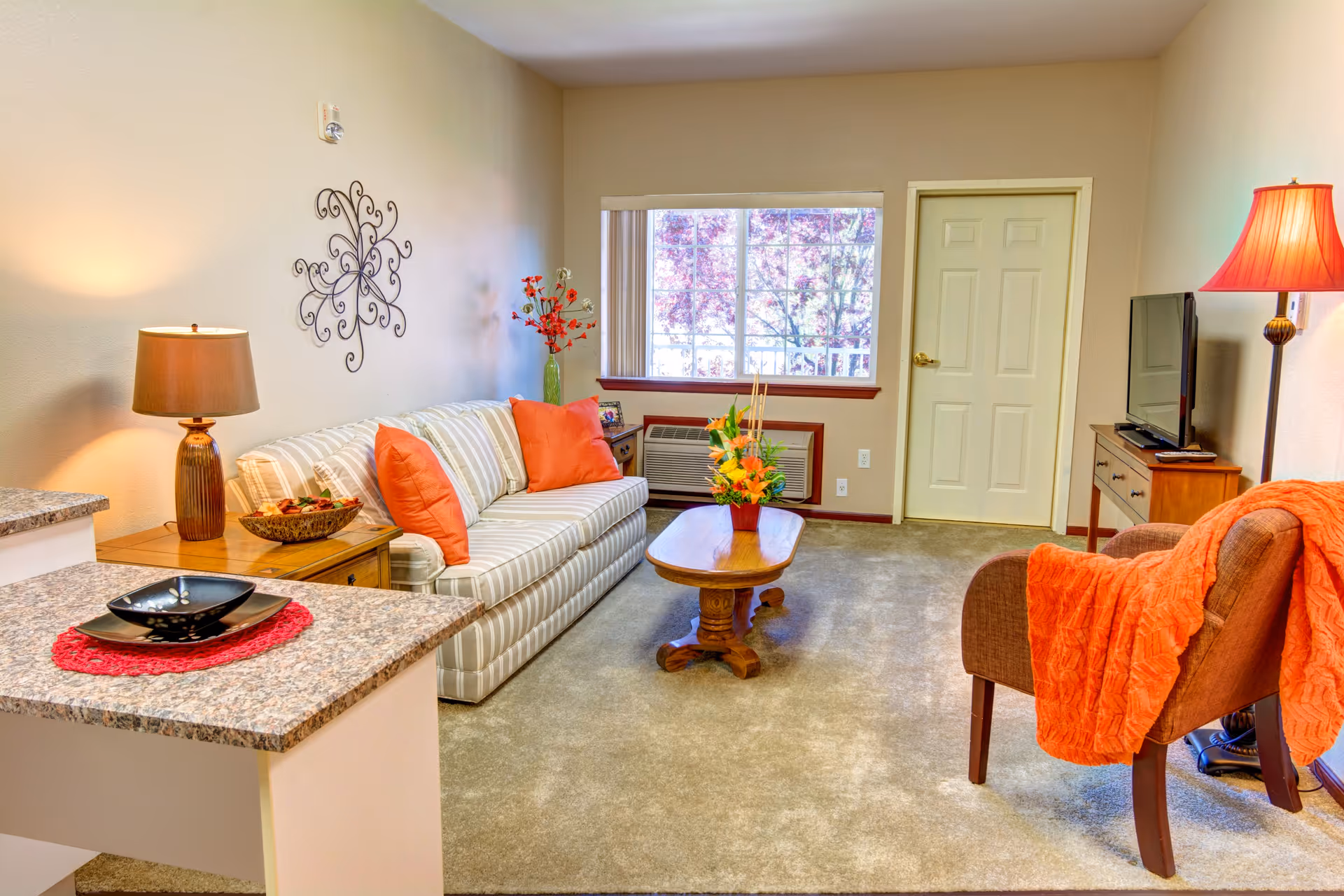 A cozy living room in a retirement community featuring a striped sofa with orange pillows, a wooden coffee table with a flower arrangement, a brown armchair with an orange throw blanket, a floor lamp with an orange shade, a TV on a wooden stand, and a window with a view of trees outside. The room has beige walls and carpeted flooring, with a small kitchen counter visible in the foreground.