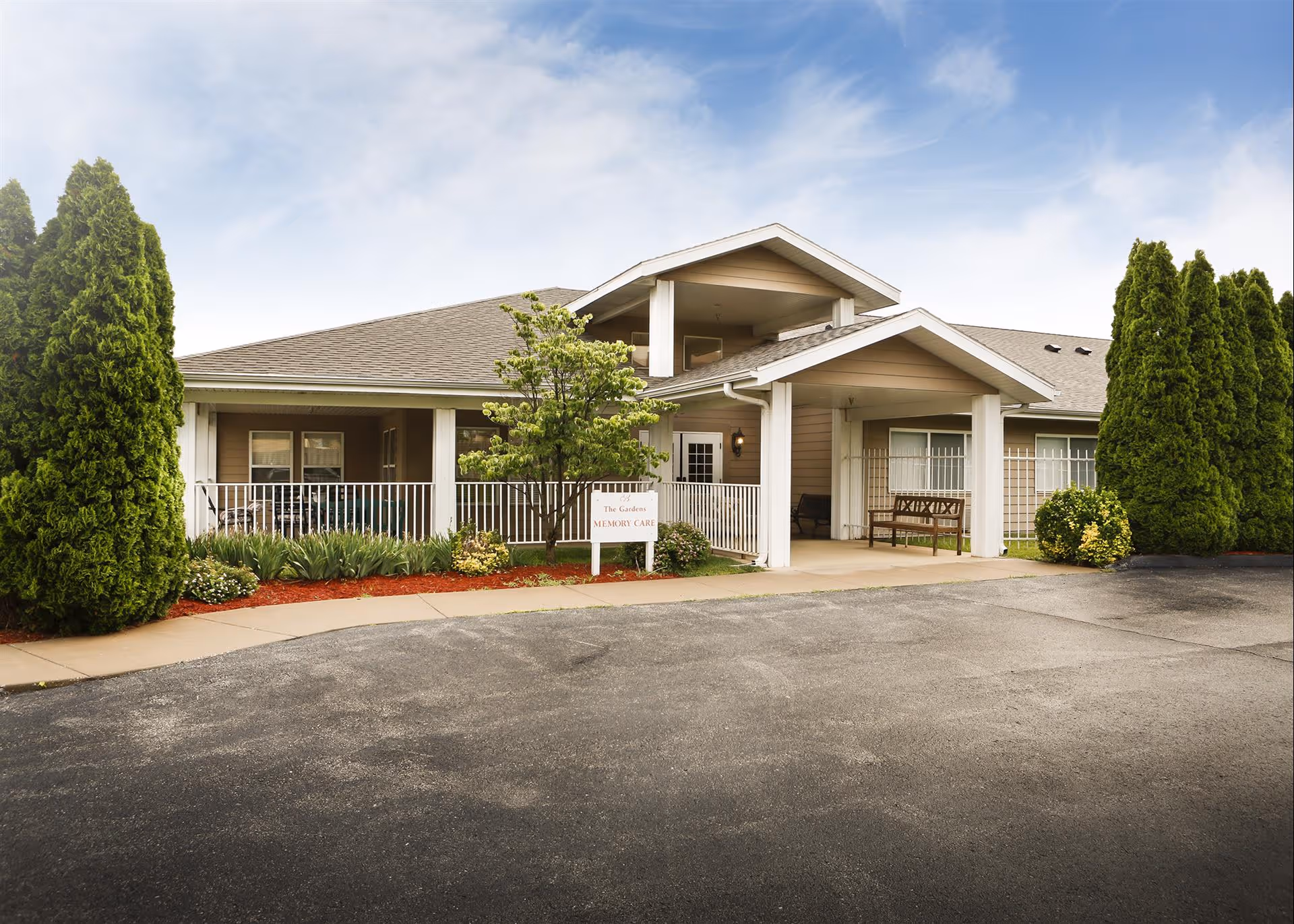 Exterior front view of The Gardens Assisted Living and Memory Care facility showing a single-story building with a covered entrance, surrounded by neatly trimmed bushes and trees under a partly cloudy sky.