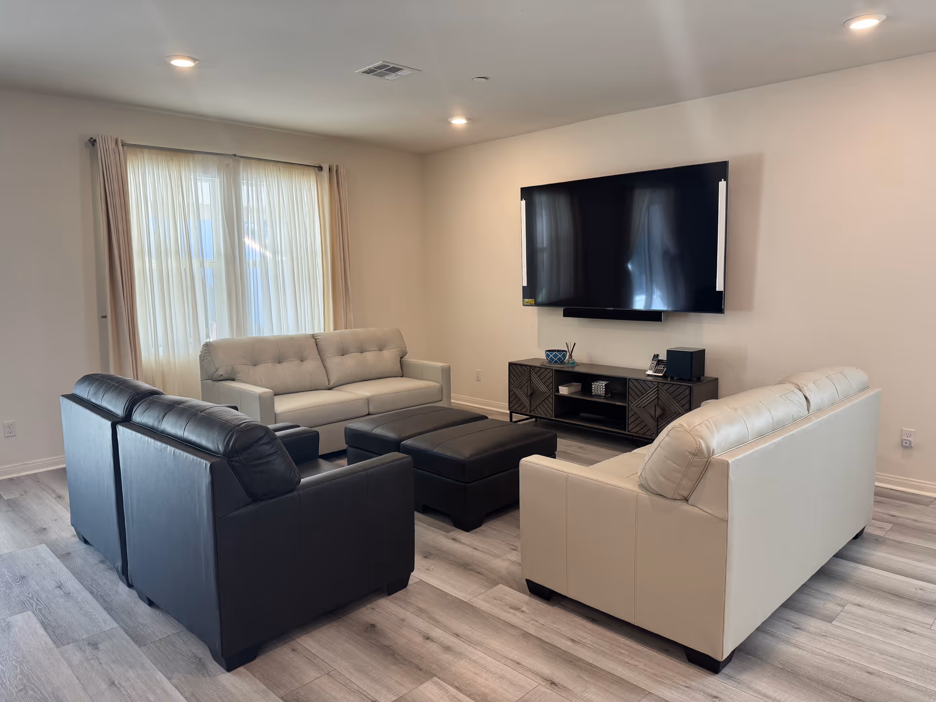 A modern living room with light and dark sofas arranged around a central ottoman facing a wall-mounted TV and media console.