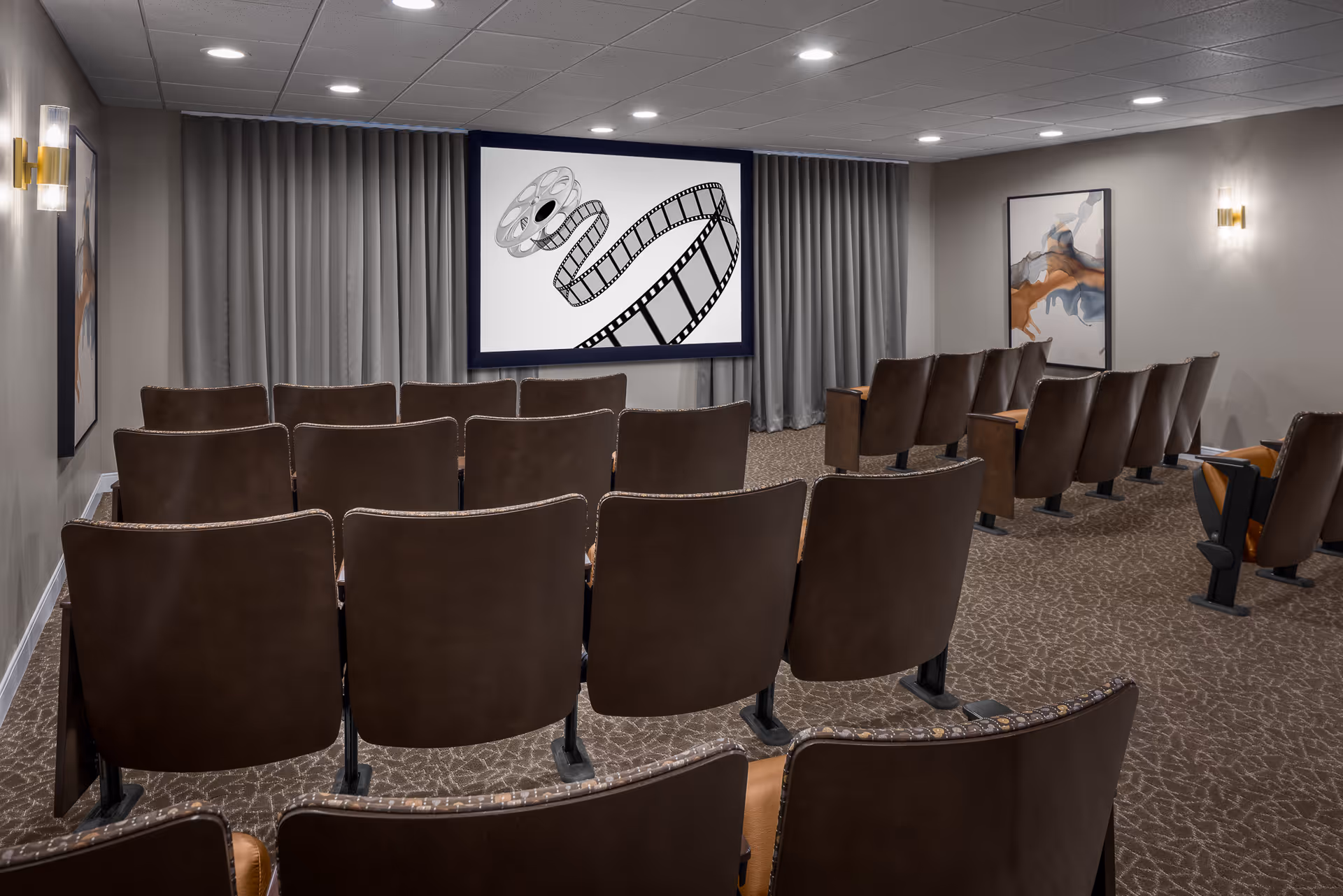 A small theater room with several rows of brown cushioned seats facing a large screen displaying an image of a film reel and filmstrip. The room has gray walls, carpeted floor, modern wall sconces, and abstract artwork on the walls.
