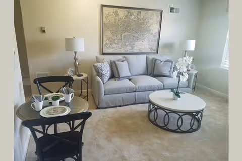 A cozy living room with a light gray sofa adorned with multiple cushions, a round coffee table with a decorative white orchid, two side tables each with a lamp, a small round dining table set for two with black chairs, and a large framed map hanging on the wall.