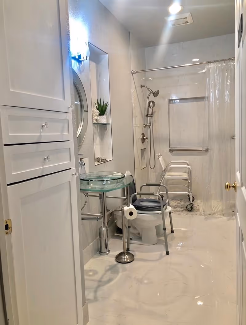 A clean, accessible bathroom with white walls and marble flooring. It features a glass sink with a round mirror above it, a toilet with a raised seat and armrests, a toilet paper holder, and a shower area with a handheld showerhead, grab bars, and a shower chair. There is also a white cabinet on the left side and a small plant on a glass shelf.