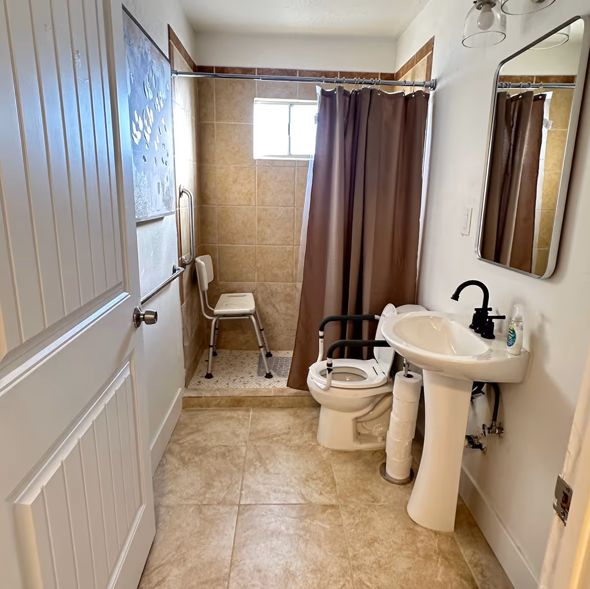 A bathroom with beige tiled floor and walls, featuring a walk-in shower with a brown curtain and a white shower chair inside. There is a white toilet with black safety rails and a pedestal sink with a black faucet. A mirror is mounted on the wall above the sink, and a stack of toilet paper rolls is placed next to the toilet. A white door is partially open on the left side of the image.