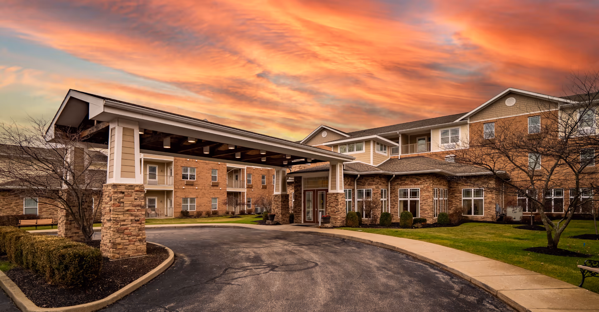 Exterior view of a senior living facility building with a covered entrance driveway, brick and siding facade, and a vibrant sunset sky in the background. The building is surrounded by neatly trimmed bushes, trees, and a paved driveway.