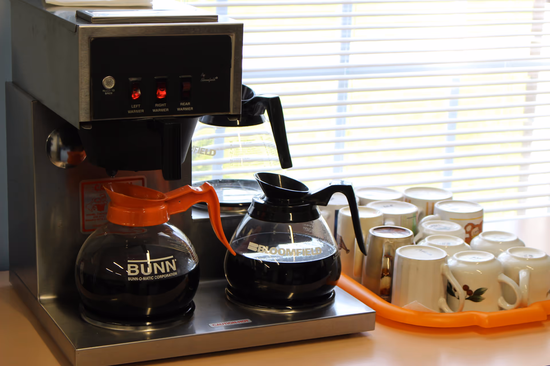 A coffee maker with two glass coffee pots, one labeled Bunn with an orange handle and the other labeled Bloomfield with a black handle, sitting on a counter next to a tray holding multiple upside-down coffee mugs. A window with blinds is in the background.