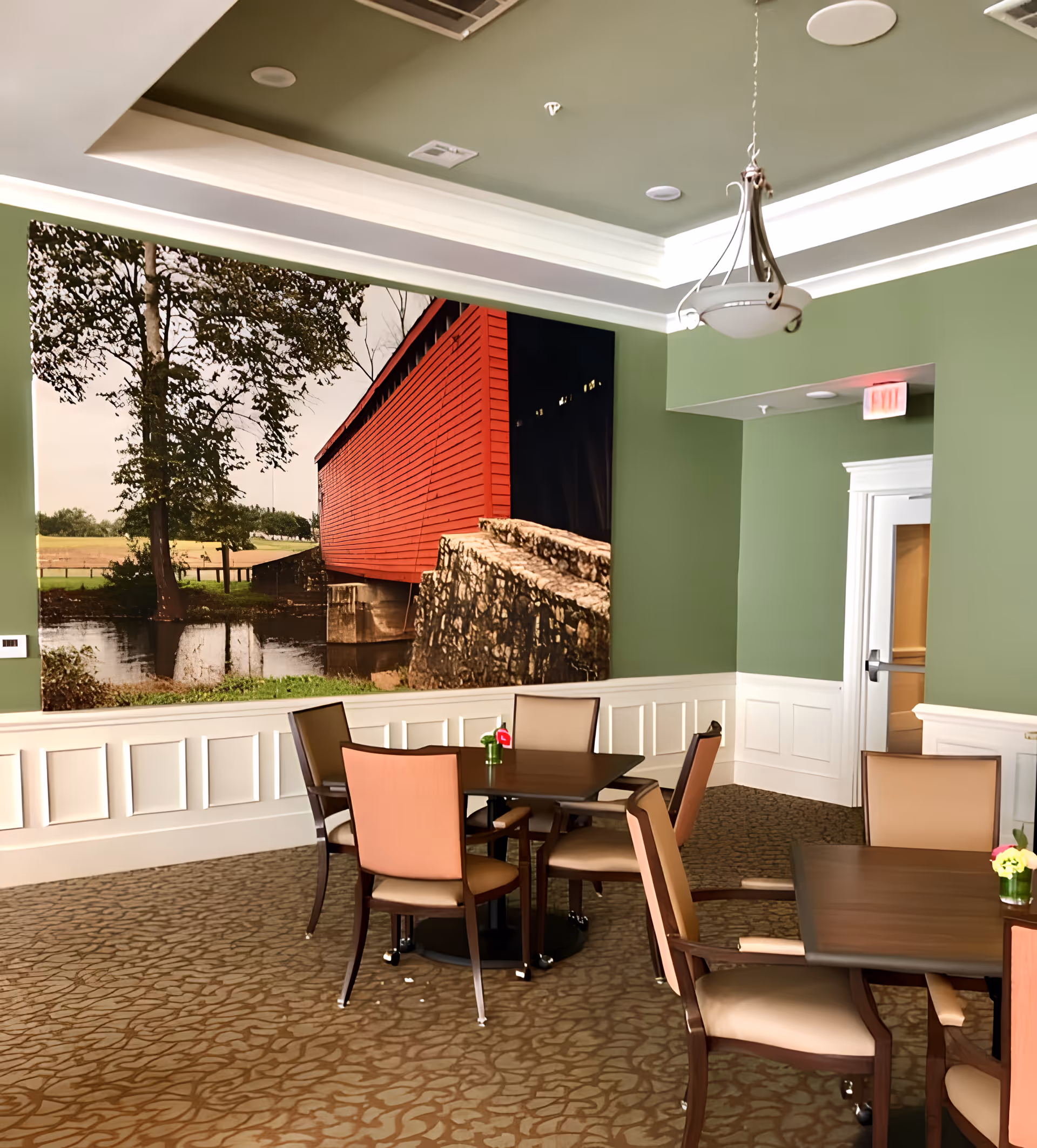 Dining area with tables and chairs in a green-walled room featuring a large mural of a red covered bridge.