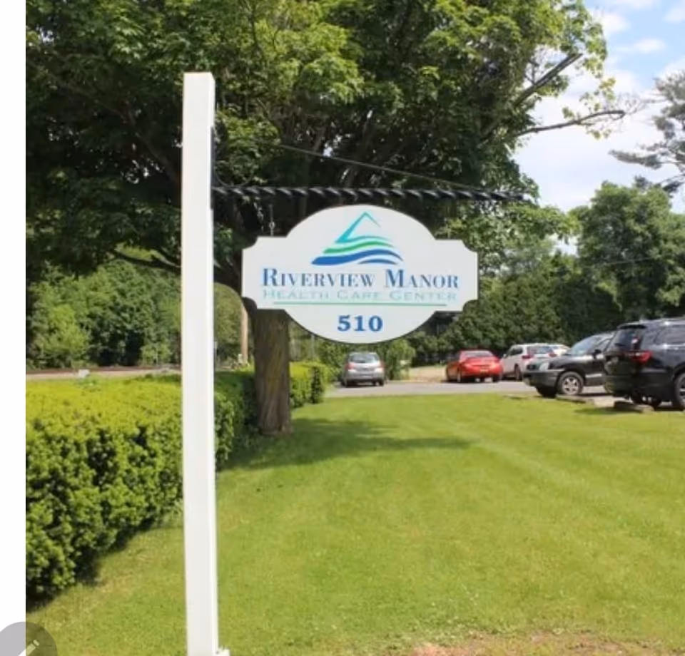 Outdoor view of a white hanging signpost with the sign reading 'Riverview Manor Health Care Center 510' surrounded by green grass, trees, and parked cars in the background under a partly cloudy sky.