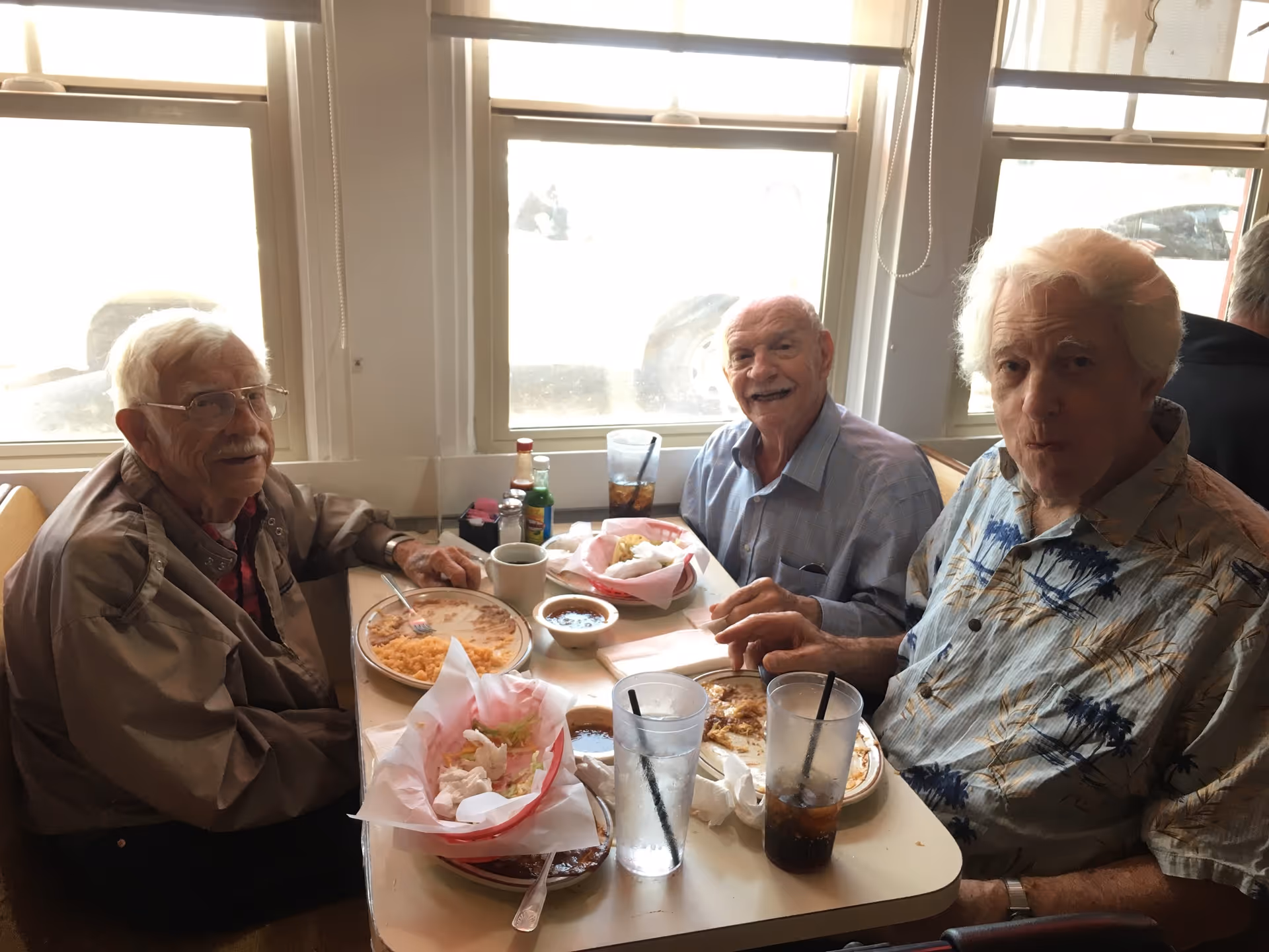 Three elderly men seated at a small dining table by windows enjoying a meal.