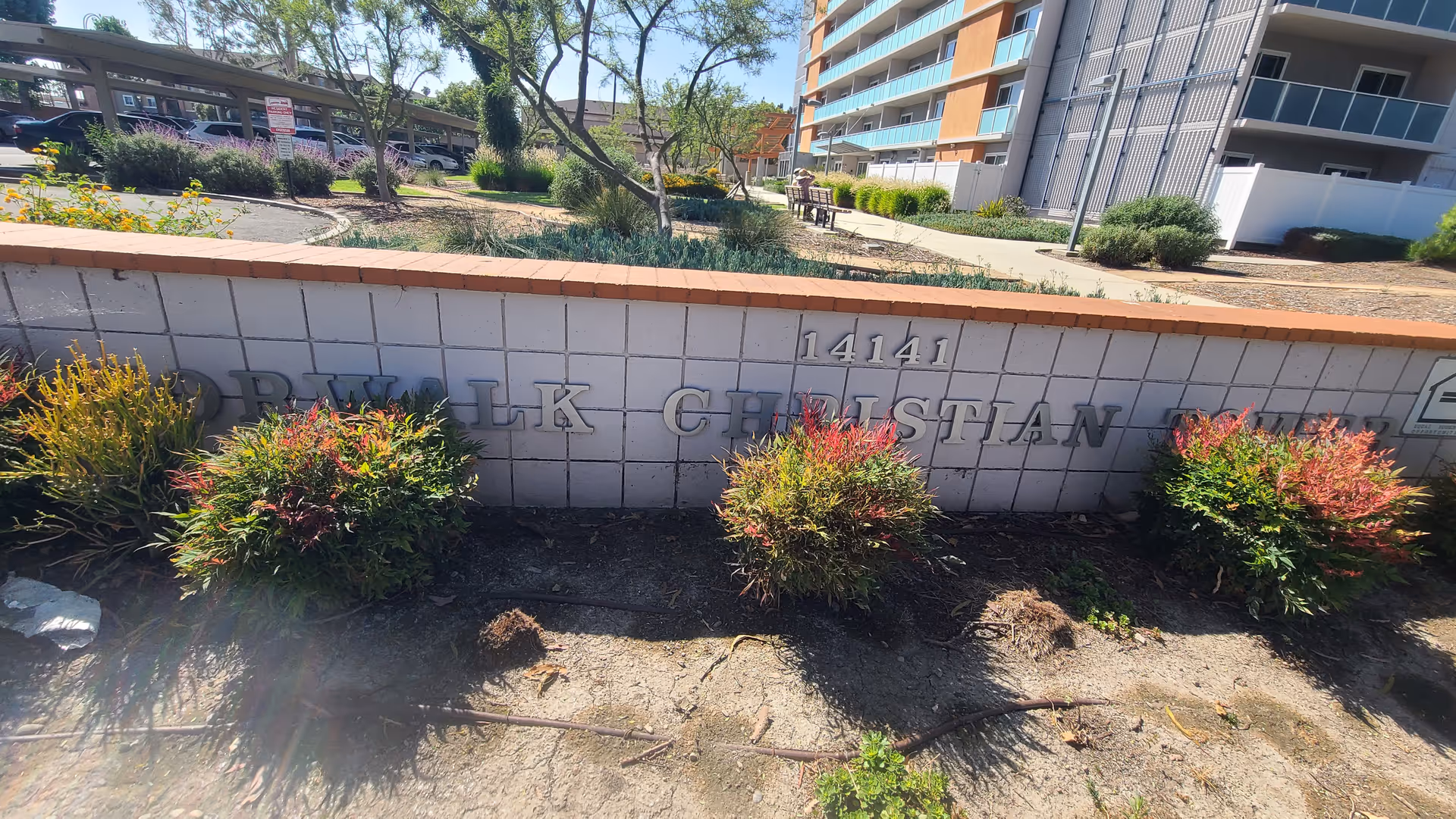 Low brick entrance wall displaying the address '14141' and part of the building name, with shrubs in front and a multi-story apartment building behind.