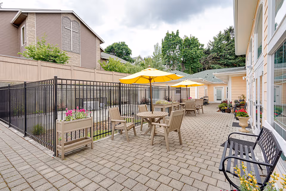 Outdoor patio area at Tabor Crest Memory Care with beige tables and chairs under yellow umbrellas, black metal bench, potted plants, and a fenced section. The area is paved with stone tiles and surrounded by buildings and trees.