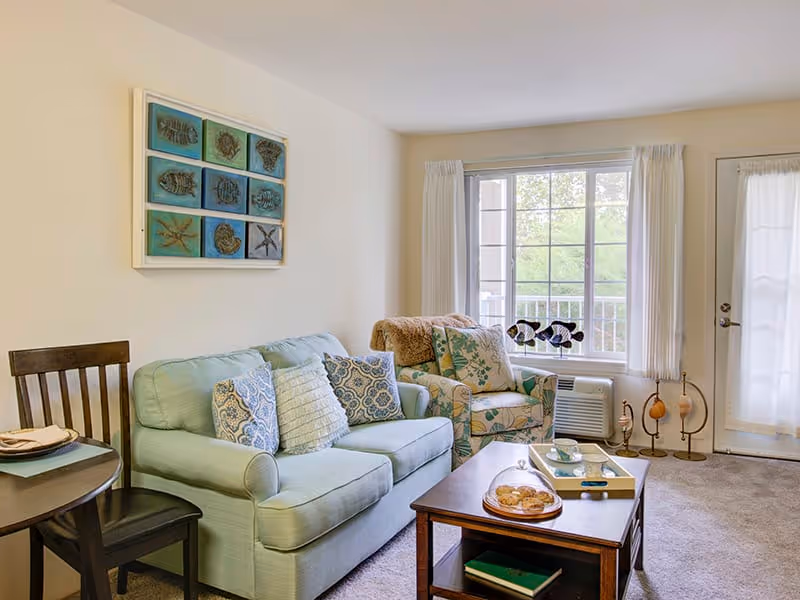 Cozy living room with a light green sofa, patterned armchair, coffee table, dining chair, and a large window/door leading to a balcony.