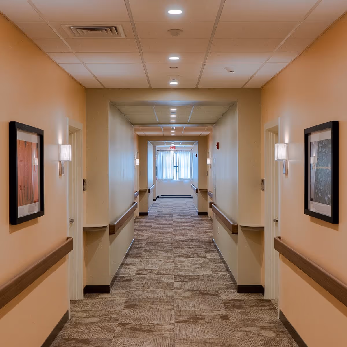A long, well-lit hallway in a senior living facility with beige walls, carpeted floor, handrails on both sides, framed artwork, and wall-mounted lights. At the end of the hallway, there is a window with white curtains allowing natural light to enter.