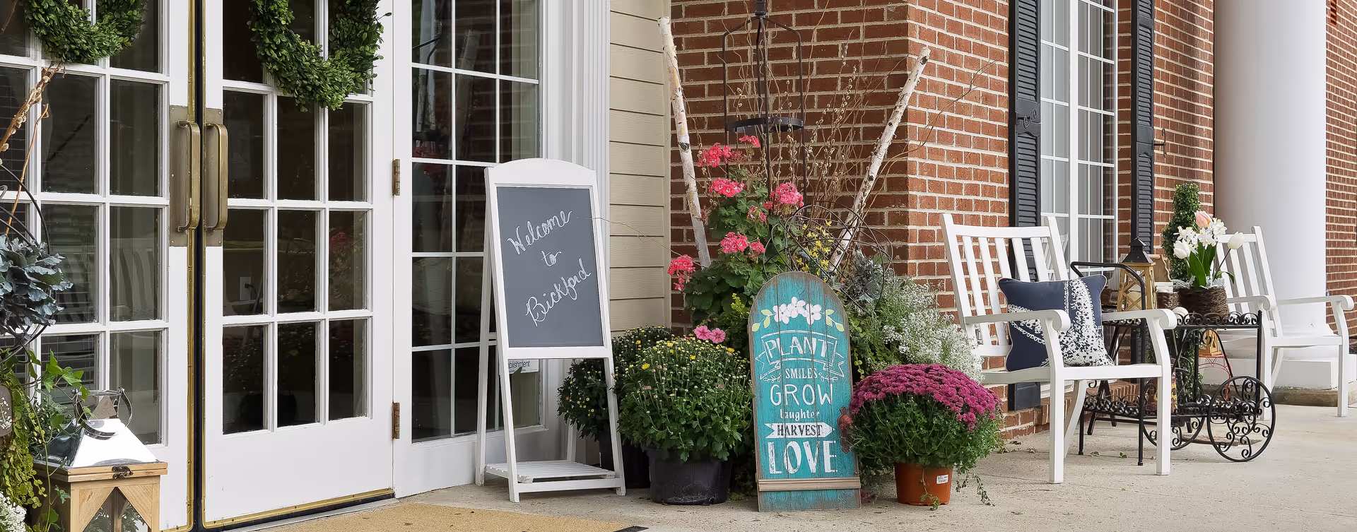 Entrance to a building with white double doors adorned with green wreaths, a chalkboard sign that reads 'Welcome to Bickford,' and a decorative sign with flowers and inspirational words. There are potted plants with pink and purple flowers, white chairs with cushions, and a small black metal table on the porch area.