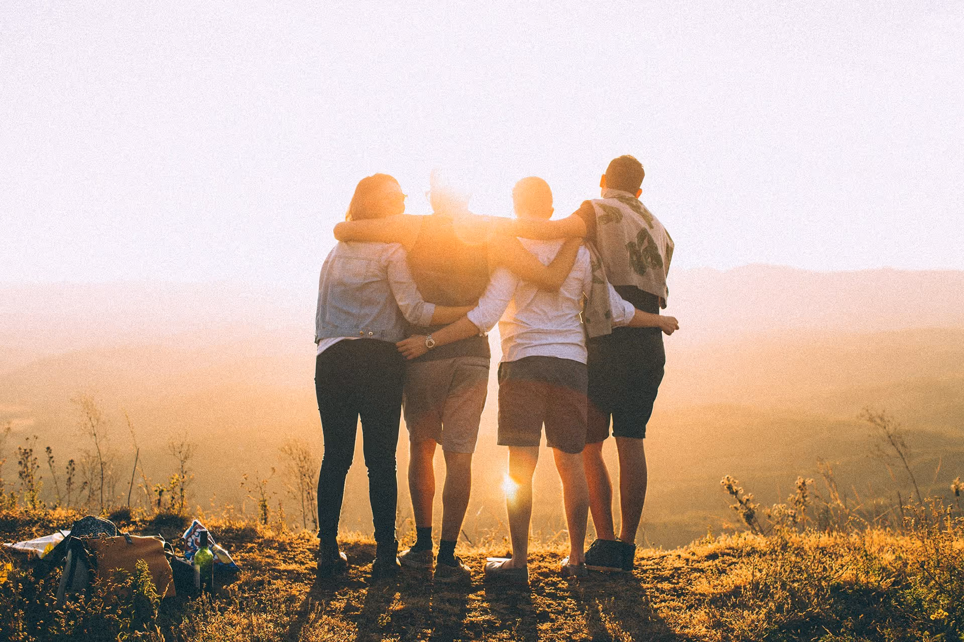 Four people standing outdoors on a grassy hill with their arms around each other, facing a bright sunset over a scenic landscape.