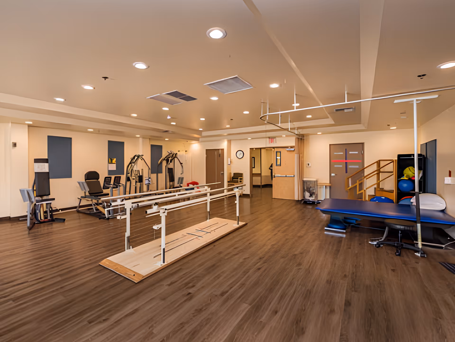 A spacious rehabilitation room with wooden flooring, parallel bars for walking exercises, exercise machines along the left wall, a blue therapy table on the right, and various exercise balls and equipment in the corner. The room is well-lit with ceiling lights and has beige walls with blue acoustic panels.