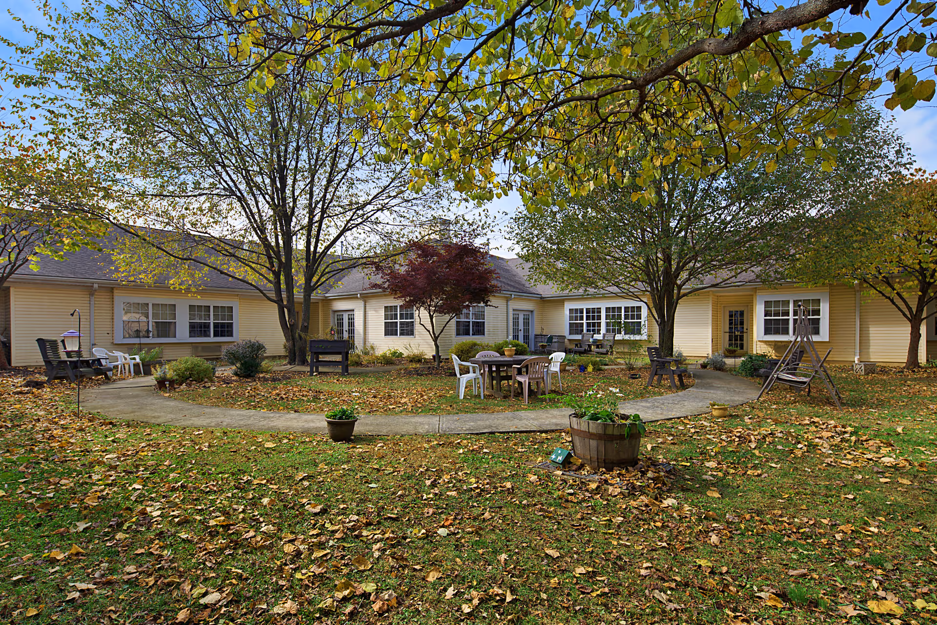 A leafy courtyard of a single-story care facility with trees, a circular concrete path, patio tables and chairs, and fallen autumn leaves.
