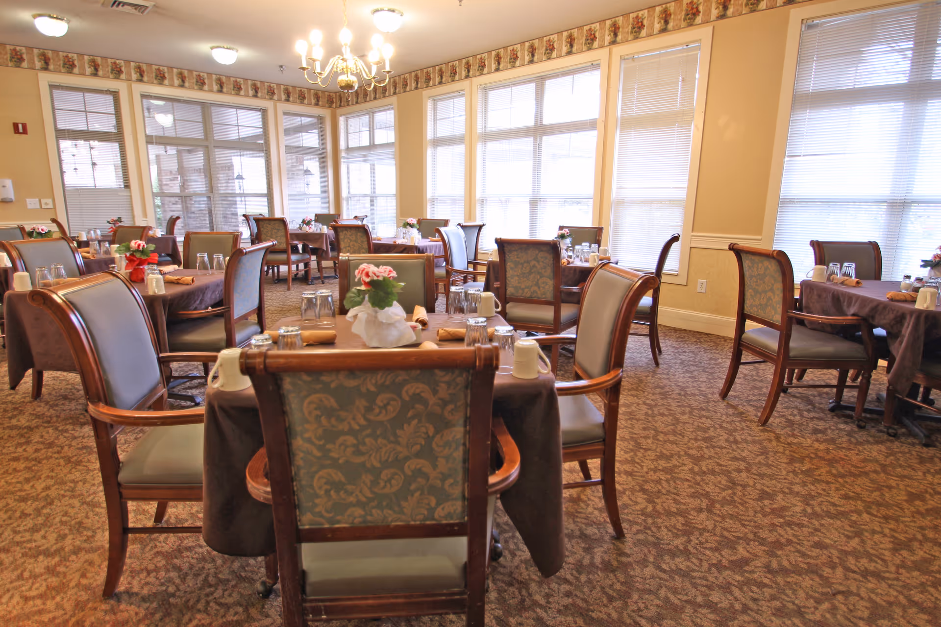 A bright dining room with multiple tables covered in brown tablecloths, each set with glasses, napkins, and small flower arrangements. The room has large windows with blinds, patterned carpet, and a chandelier hanging from the ceiling.