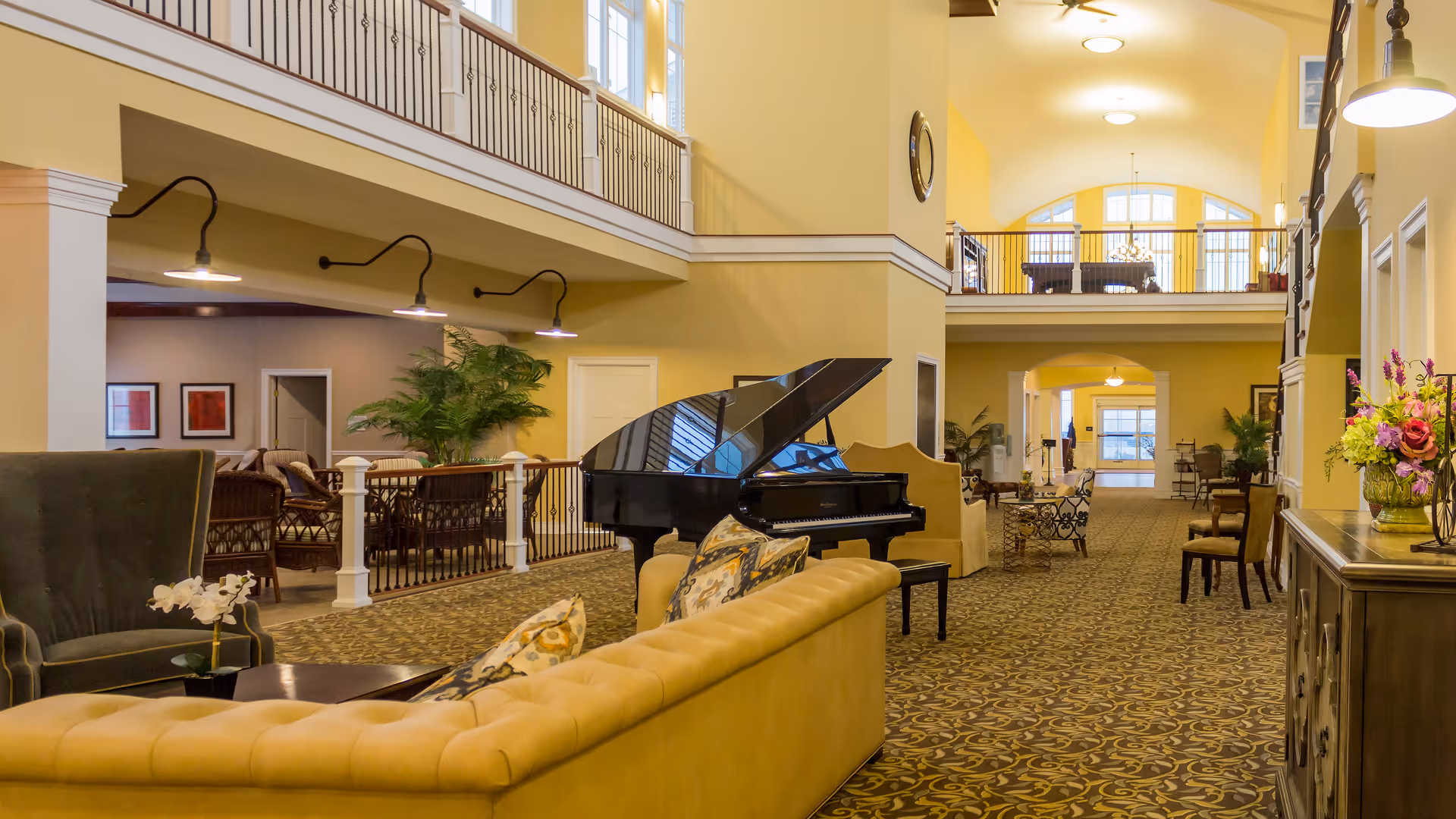 Spacious and elegant senior living facility common area with a grand piano, comfortable seating including a yellow tufted sofa and armchairs, decorative pillows, patterned carpet, and warm yellow walls. The area features a high ceiling with a balcony railing above, large windows letting in natural light, and various plants and flowers adding a touch of greenery.