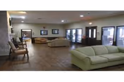A spacious living room area with wooden flooring, multiple beige and light green sofas arranged around the room, several chairs along the left wall, and large windows and glass doors on the right side allowing natural light to enter.