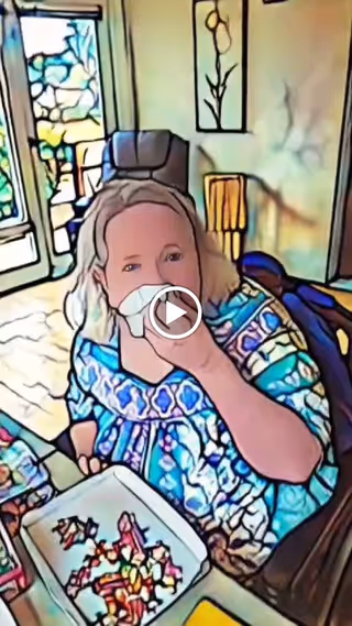 A woman sitting at a table in a room with large windows and a door leading outside. She is holding a tissue to her nose and wearing a colorful patterned top. On the table in front of her is a tray with various small items or pieces of food. The room has light-colored walls with a framed flower artwork and a lamp in the background.