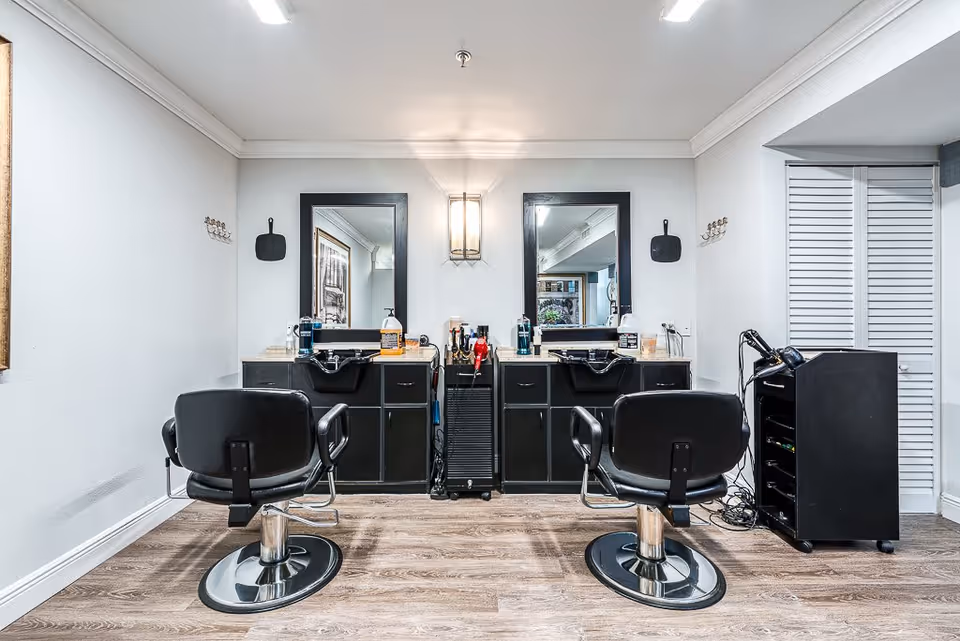 Small salon station with two black styling chairs facing mirrored sinks and countertops stocked with hair products.