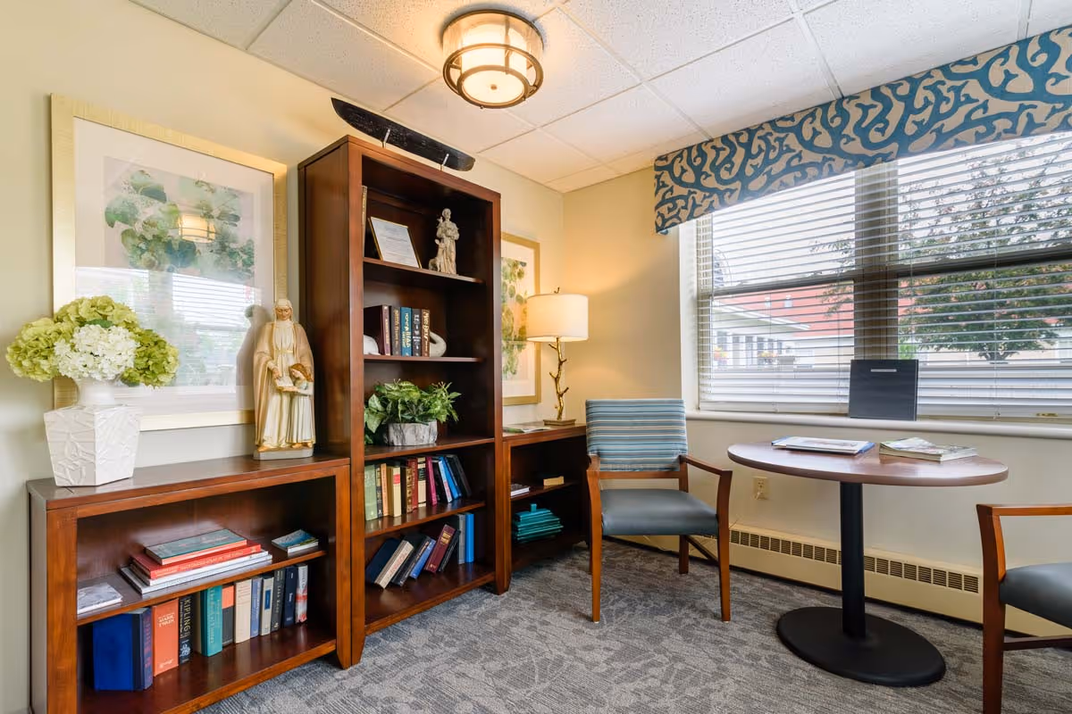 Bright seating and reading nook with wooden bookshelves, two chairs, a round table, lamp, and a large window with blinds.