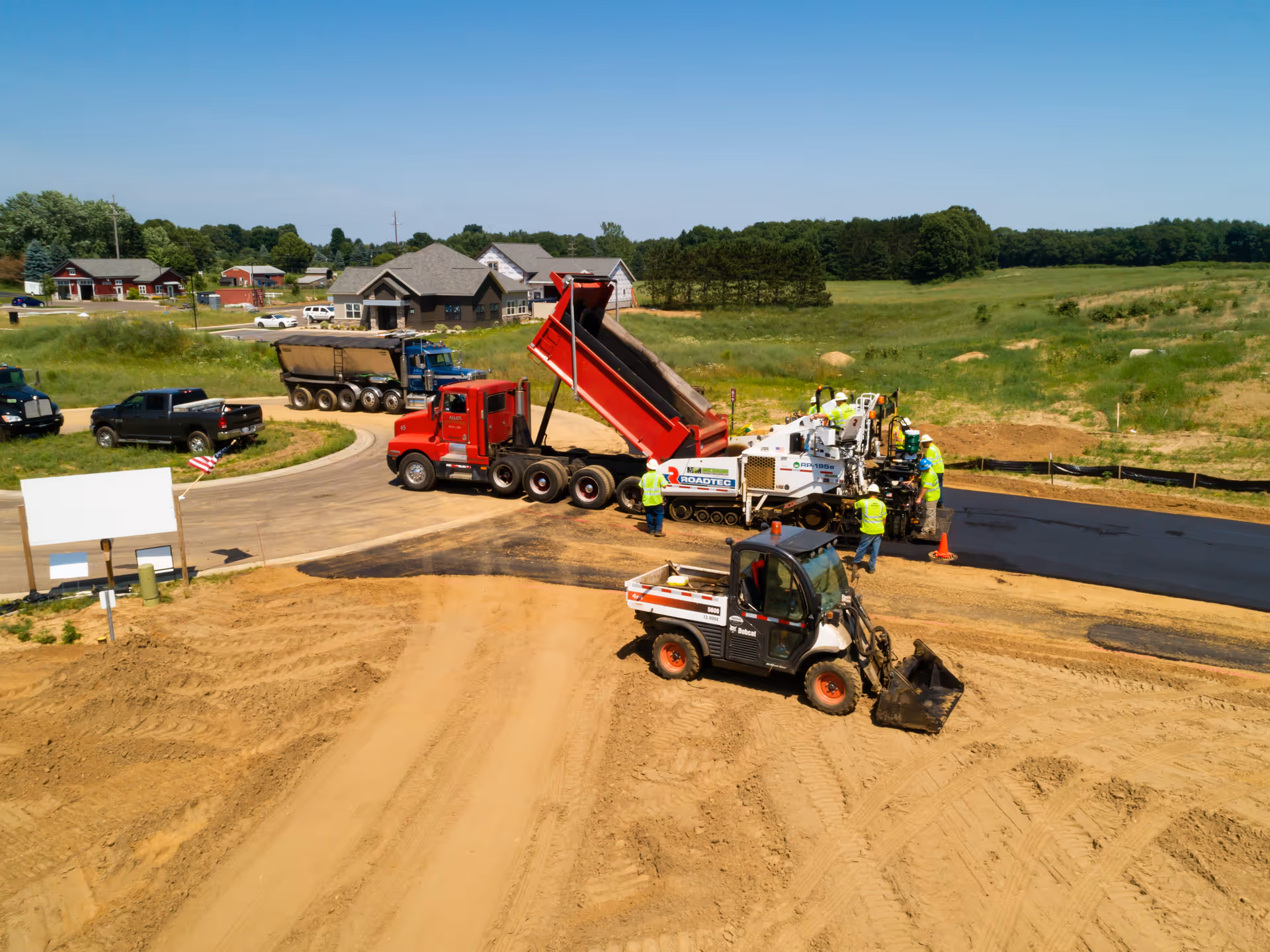 Construction site with workers paving a road using heavy machinery including a red dump truck unloading asphalt and a white paving machine. Several vehicles and houses are visible in the background under a clear blue sky.