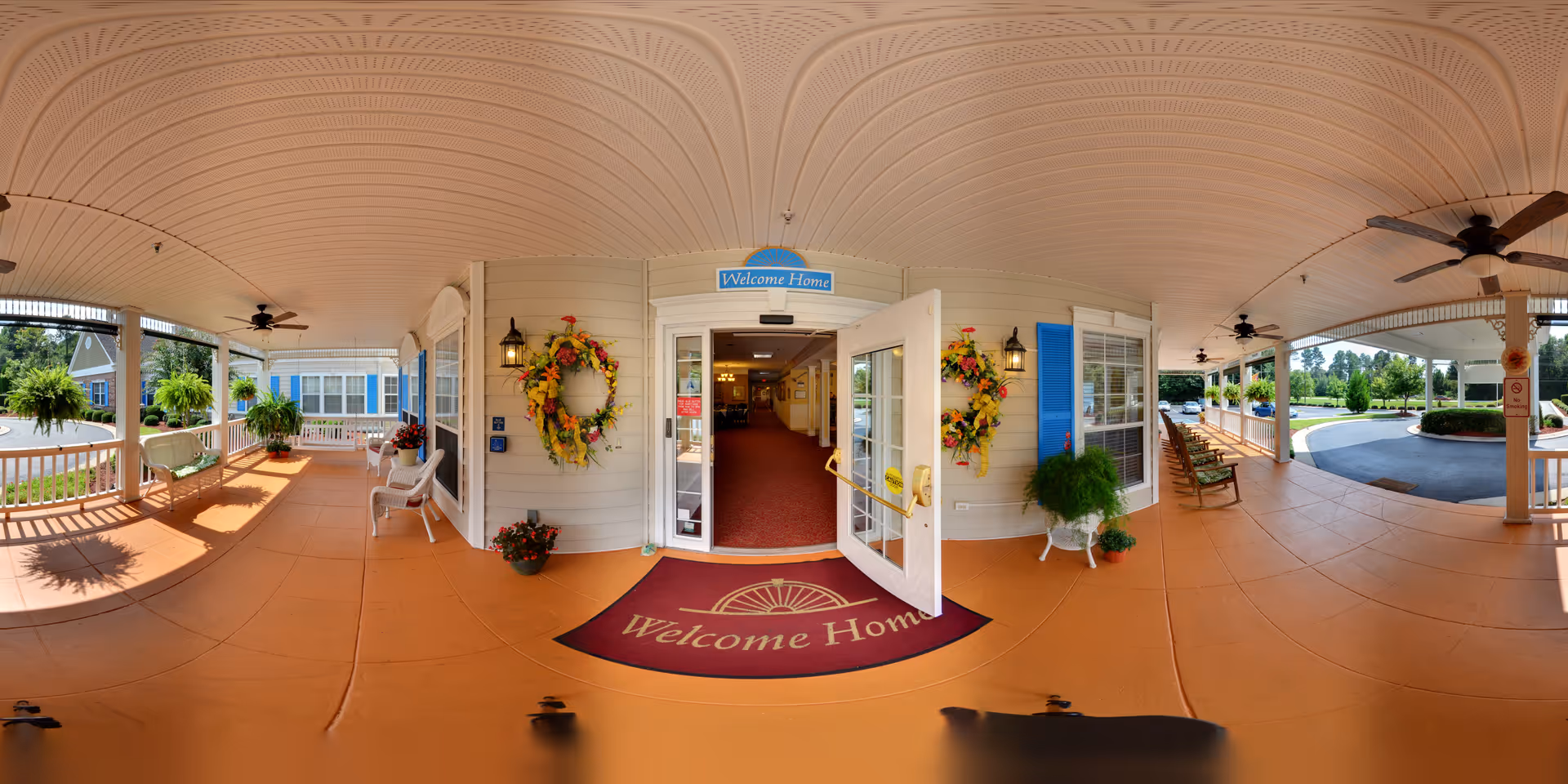 Wide panoramic view of a covered porch entrance to a senior living facility with a 'Welcome Home' mat and sign above the door. The porch features ceiling fans, hanging plants, rocking chairs, and decorative wreaths on the walls. The entrance door is open, showing a hallway inside the building. The outdoor area includes a driveway and landscaped greenery.