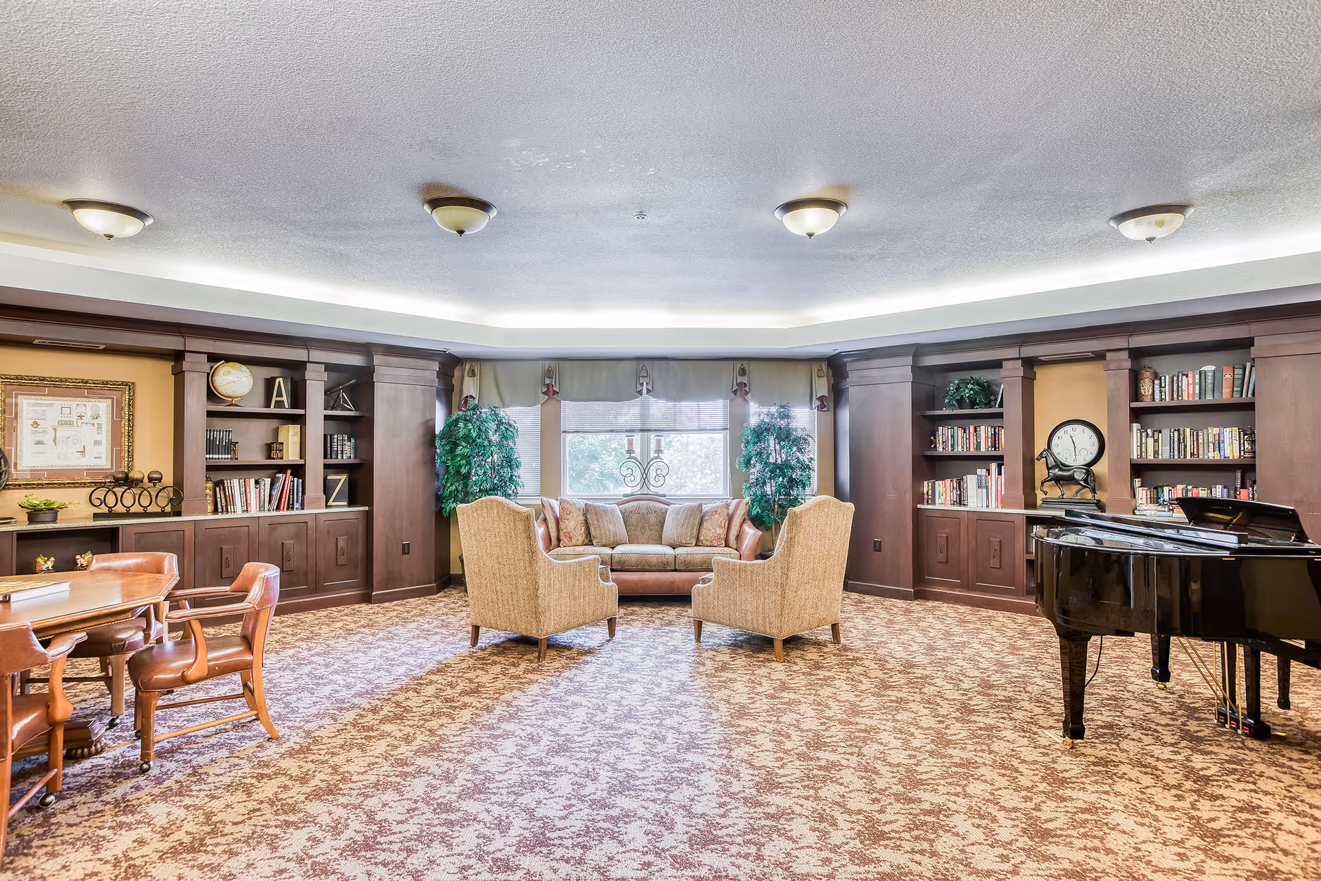 A spacious and well-lit common area with patterned carpet flooring, two beige armchairs facing a sofa near a large window with curtains, flanked by two tall green plants. The room features built-in dark wood bookshelves filled with books and decorative items, a round wooden table with four leather chairs on the left, and a black grand piano on the right. The ceiling has multiple light fixtures and recessed lighting around the perimeter.