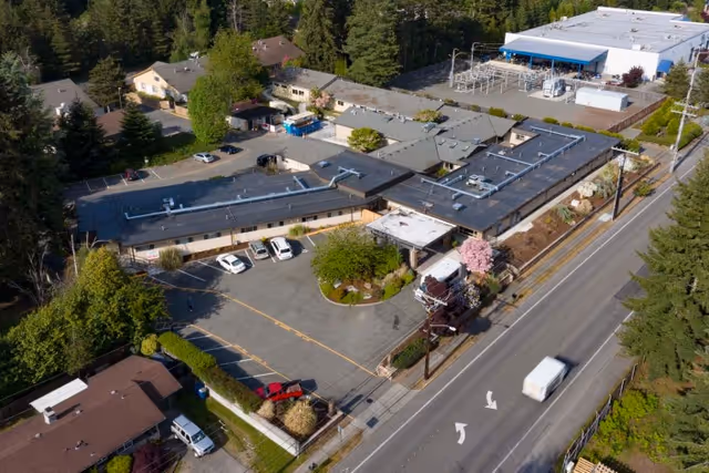 Aerial view of Lynnwood Post Acute Rehabilitation Center showing a single-story building with a parking lot in front, surrounded by trees and nearby residential houses. A road runs alongside the facility with a white vehicle driving on it.