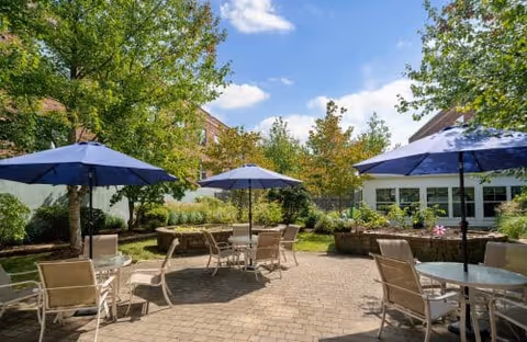 Sunlit courtyard patio with round tables, chairs, and blue umbrellas surrounded by trees and buildings.