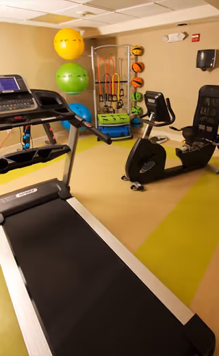 A small fitness room with exercise equipment including a treadmill, a recumbent stationary bike, colorful exercise balls, resistance bands, and medicine balls against a beige wall.