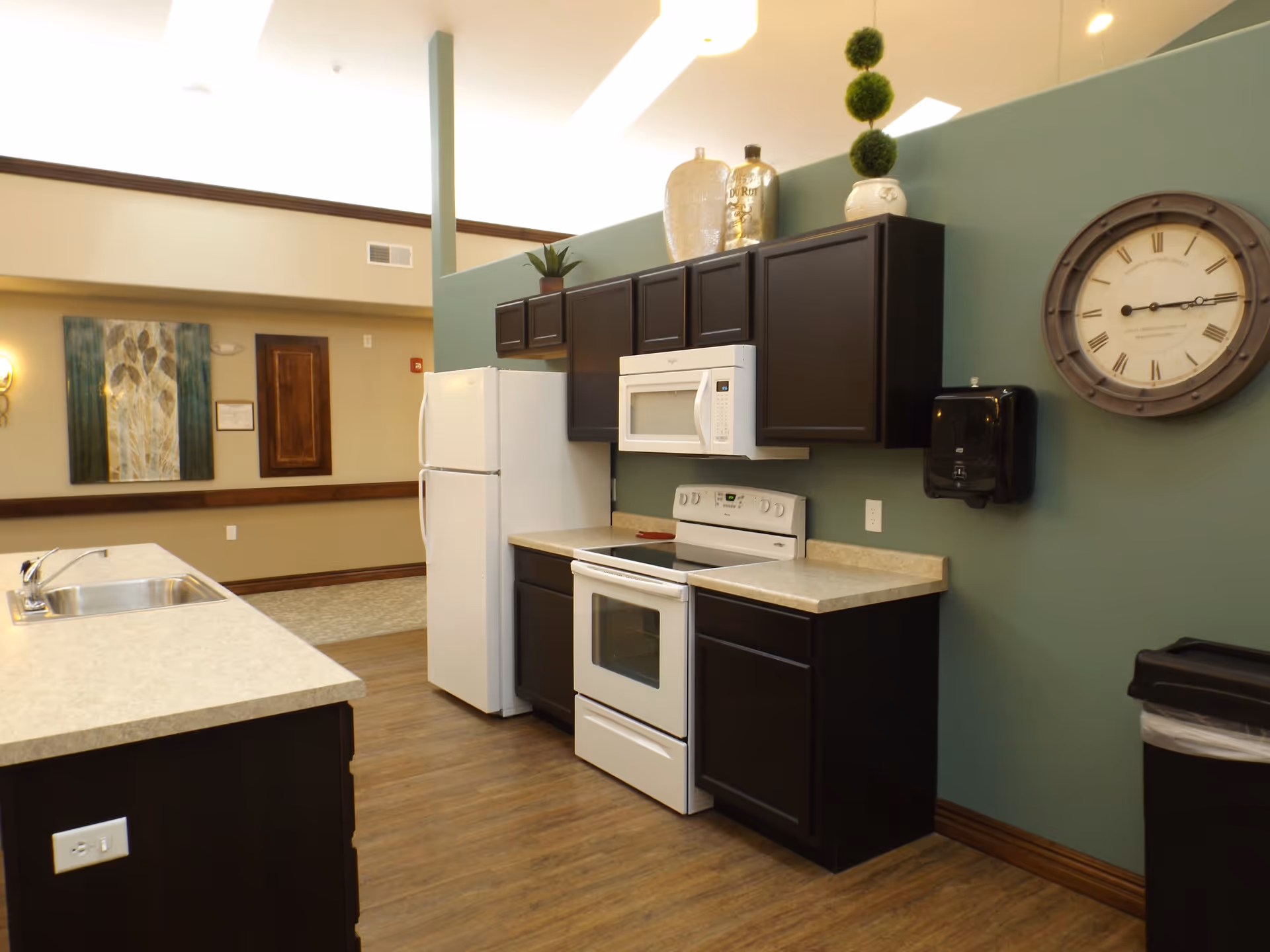Interior view of a kitchen area with dark brown cabinets, a white refrigerator, white stove with oven, and a white microwave. The kitchen has beige countertops and a sink on an island. A large round wall clock and a black paper towel dispenser are mounted on a green wall. Decorative items are placed on top of the cabinets. The floor is wood, and there is a hallway with artwork visible in the background.