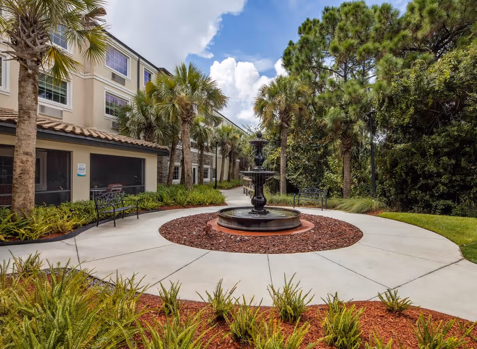 Outdoor courtyard area at Discovery Village At Stuart featuring a circular concrete pathway surrounding a black tiered water fountain. The courtyard is landscaped with palm trees, green shrubs, and red mulch. There are benches along the pathway and a building with screened windows on the left side. The sky is partly cloudy.
